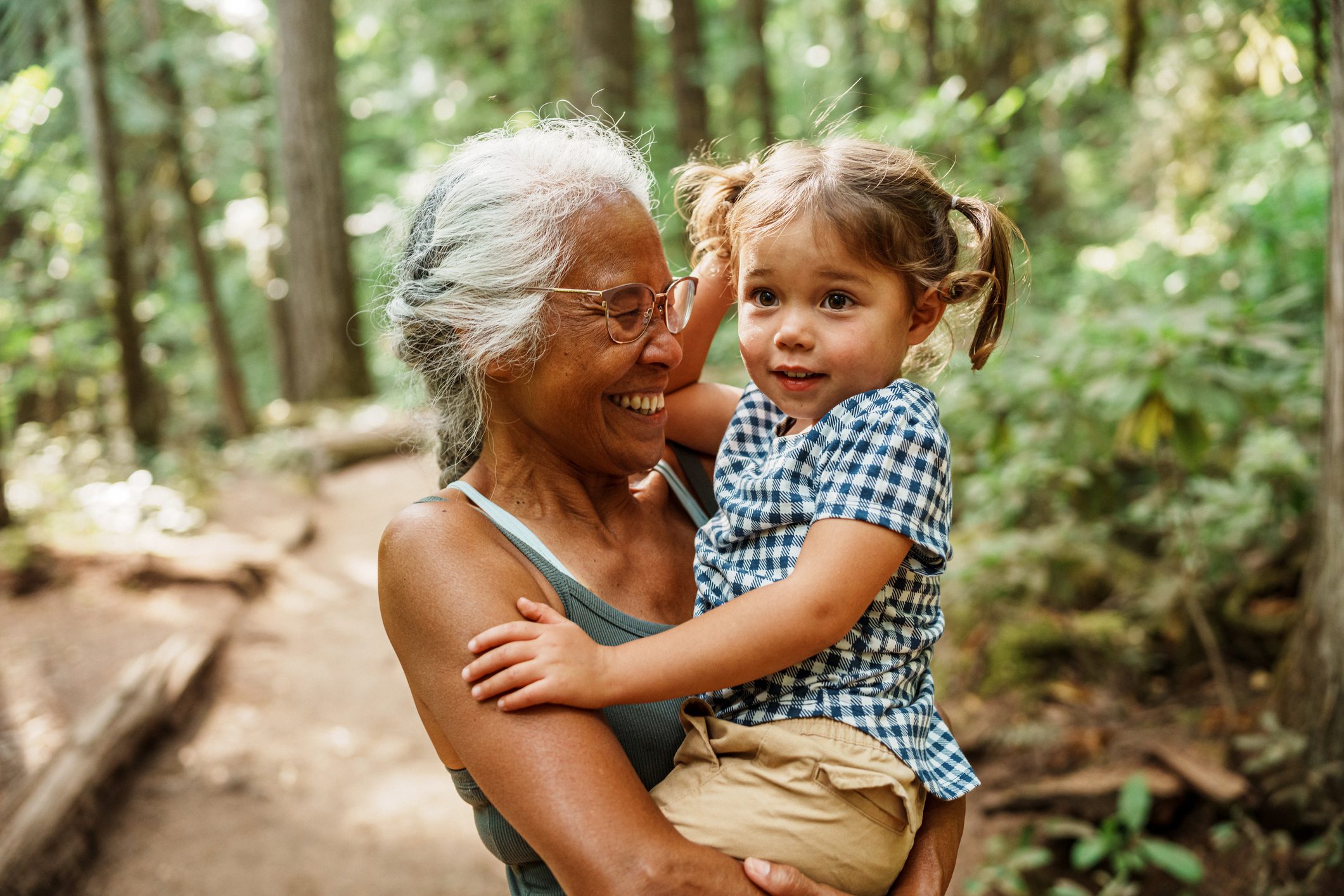 A healthy and adventurous senior woman of Hawaiian and Chinese descent laughs joyfully while carrying her adorable three year old Eurasian granddaughter during a hike through a lush forest in Oregon.