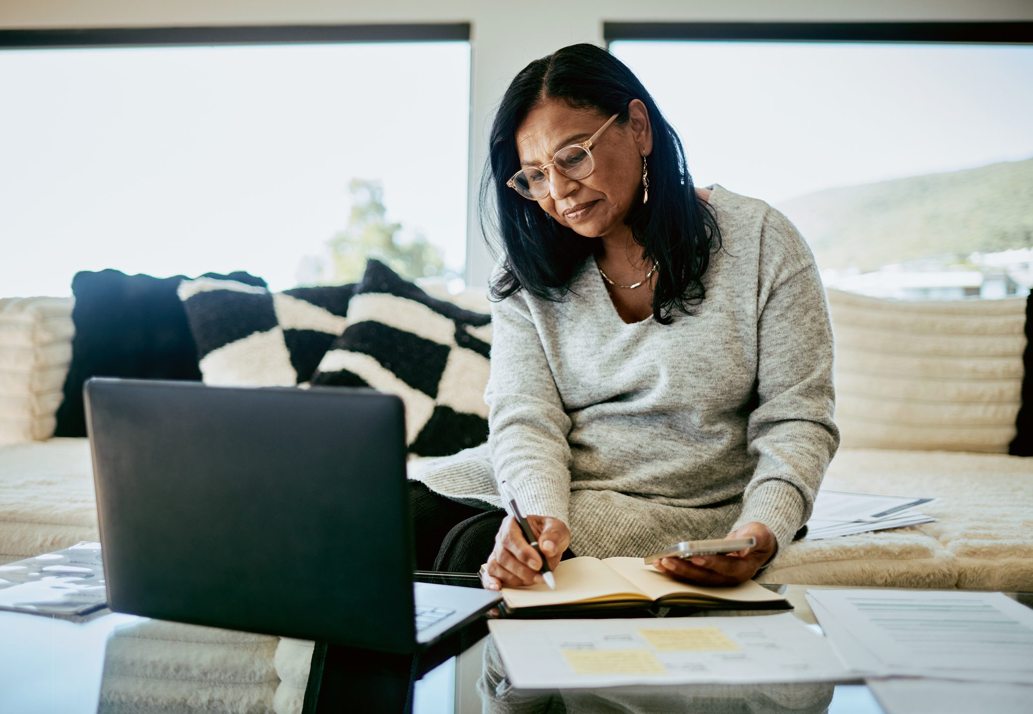 A middle-aged woman with glasses, of South Asian descent, working remotely in her living room with a laptop, notebook, and smartphone.