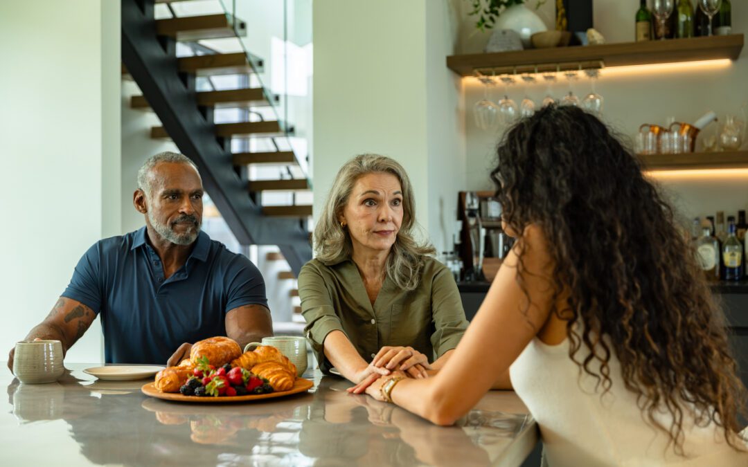 A mature adult couple sits at a modern dining table in the morning, engaged in a serious conversation with their daughter whose back is to the camera. The mother reaches across to hold her daughter's hand in a gesture of support, while the father watches with a concerned expression, a coffee mug in front of him. A plate of croissants and fresh berries sits on the polished counter, with an open staircase and shelves of glassware visible in the contemporary home interior.