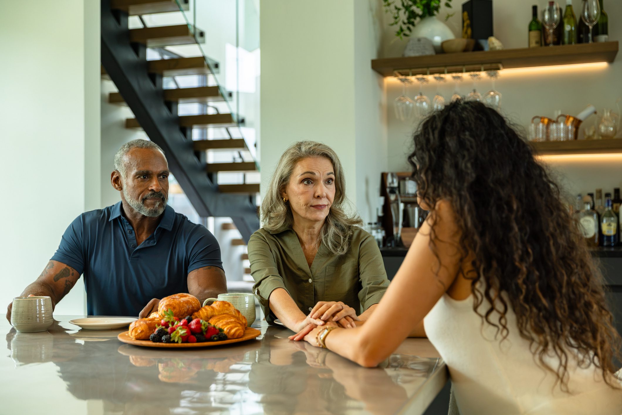 Mature Couple Comforting Adult Daughter at Kitchen Counter During Serious Family Conversation A mature adult couple sits at a modern dining table in the morning, engaged in a serious conversation with their daughter whose back is to the camera. The mother reaches across to hold her daughter's hand in a gesture of support, while the father watches with a concerned expression, a coffee mug in front of him. A plate of croissants and fresh berries sits on the polished counter, with an open staircase and shelves of glassware visible in the contemporary home interior.