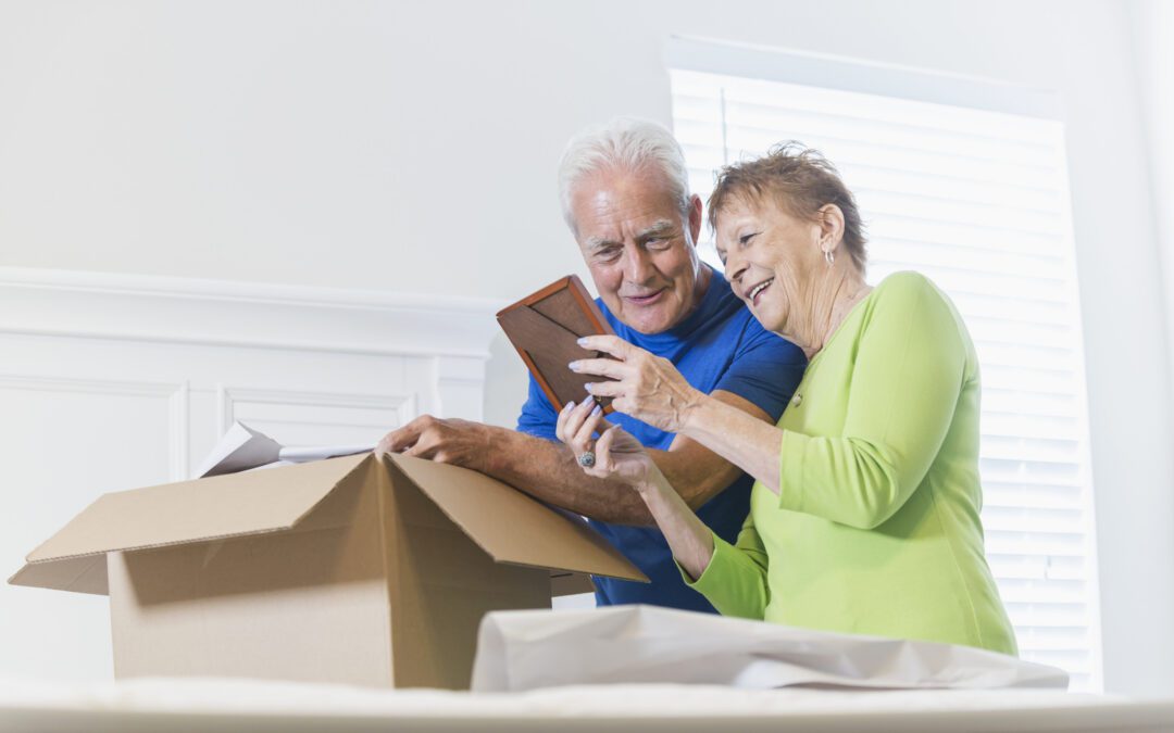 A senior couple moving house, packing a cardboard box in the bedroom. They are talking and smiling, looking at a picture frame which is bringing back memories.